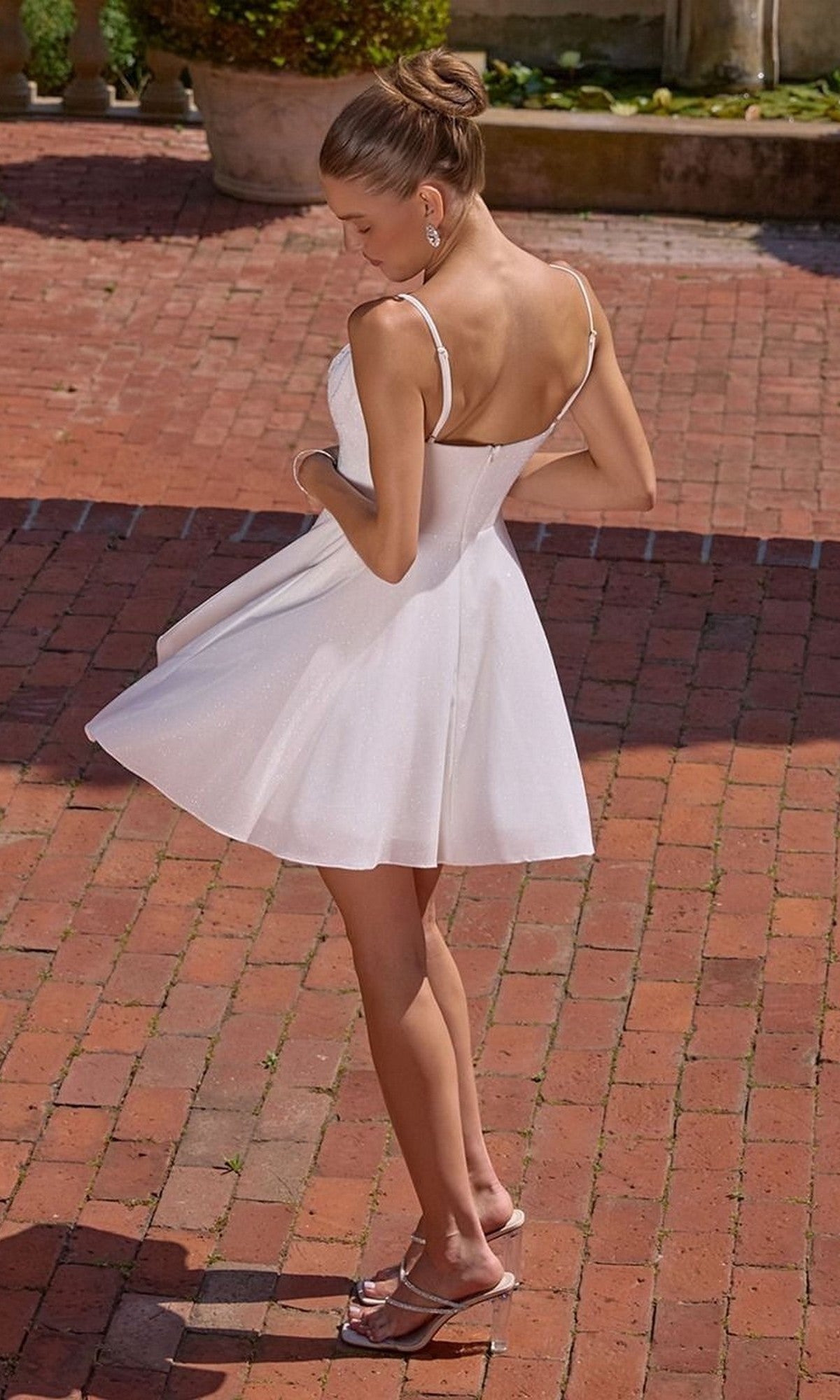 Wearing the Amoris T103W Short Homecoming Dress, a woman in a sleeveless white fit-and-flare style and heeled sandals stands on a brick patio, looking down over her shoulder with one hand on the dress.